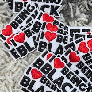 Puede incluir: Parches blancos con el texto "I ❤️ BEING BLACK" en letras negras y un corazón rojo. Los parches están esparcidos sobre una superficie gris texturizada.