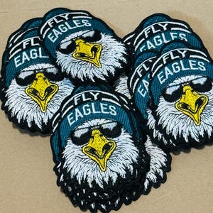 May include: Embroidered patches featuring a stylized eagle wearing sunglasses and a teal beanie with the text "FLY EAGLES". The eagle has a yellow beak and is set against a white background. Patches are stacked on a brown surface.