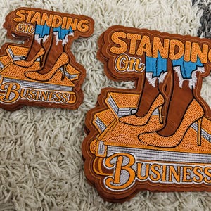Puede incluir: Parche bordado con las palabras "STANDING ON BUSINESS" en naranja y blanco. El parche presenta un par de botas de tacón alto marrones con vaqueros azules, de pie sobre una pila de libros. El parche tiene un borde marrón.