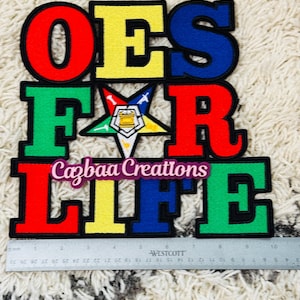 以下が含まれることがあります： 赤、黄、青、緑で「OES FOR LIFE」と書かれたカラフルな刺繍パッチ。中央には星のエンブレムがあります。「Cazbaa Creations」の文字も表示されています。パッチの長さは約13cmです。