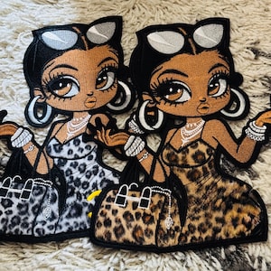 May include: Two embroidered patches featuring stylized figures with dark skin, large eyes, and sunglasses perched on their heads. Each figure wears a leopard print dress, layered necklaces, hoop earrings, and bracelets, while holding a matching handbag.
