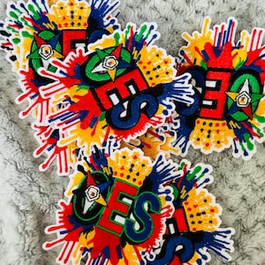 May include: Embroidered patches with the letters "E S" in red, green, and blue, topped with a gold crown. The patches feature a colorful splatter design in red, yellow, blue, and green.