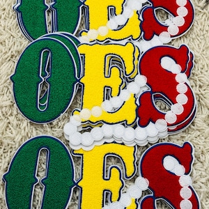 Pode incluir: Três conjuntos de emblemas de chenille que formam a palavra OES, com um O verde, um E amarelo e um S vermelho. Cada conjunto é acentuado com um design de cordão branco bordado semelhante a pérolas que se curva sobre as letras.
