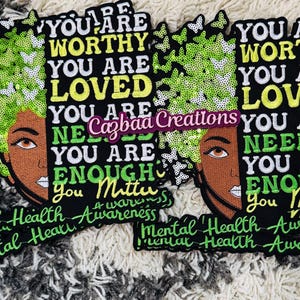Könnte beinhalten: Zwei schwarze Aufnäher mit gesticktem Text und dem Gesicht einer Frau. Der Text lautet: "YOU ARE WORTHY, YOU ARE LOVED, YOU ARE NEEDED, YOU ARE ENOUGH, YOU MATTER." Das Haar der Frau ist grün und mit Schmetterlingen verziert. Unten steht "Mental Health Awareness."