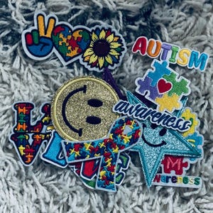 Puede incluir: Un conjunto de parches bordados coloridos con temas de concienciación sobre el autismo. Los diseños incluyen un signo de la paz, piezas de rompecabezas, un girasol, una carita sonriente, la palabra "AUTISMO" y la palabra "concienciación".