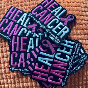 May include: A collection of black embroidered patches with the words "HEAL CANCER" in pink and light blue. Each patch includes a pink ribbon and the text "There's nothing that he cannot do, Luke 1:37."