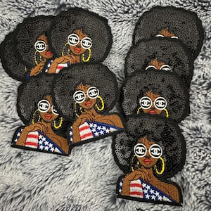 Puede incluir: Parches bordados que representan a una mujer negra con afro, gafas de sol y aros dorados. La mujer lleva una camiseta con un diseño de bandera americana roja, blanca y azul. Los parches están sobre una superficie gris y difusa.