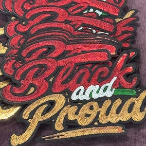 Peut inclure: Une pile d'écussons brodés ornés de sequins rouges, blancs et dorés. Le texte indique Black and Proud dans une écriture cursive. Chaque écusson possède une bordure en tissu noir et ils sont superposés, mettant en valeur la texture brillante.