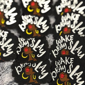 Może przedstawiać: Czarne, haftowane naszywki z białym napisem "WAKE PRAY SLAY". Każda naszywka przedstawia stylizowaną ilustrację kobiety o brązowej skórze, czerwonych ustach i złotych kolczykach w kształcie obręczy. Naszywki są zgrupowane.