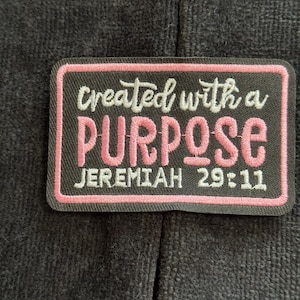 May include: A rectangular patch with a black background and a pink border. The patch features the words "created with a PURPOSE" in white and pink script, with "JEREMIAH 29:11" in white block letters.