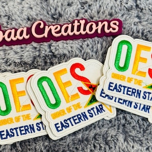 May include: Multiple embroidered patches featuring the text OES and Order of the Eastern Star in green, yellow, and red lettering. Each patch includes a yellow star emblem with a central symbol. A dark purple sign reads Cazbaa Creations.