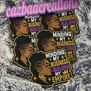 May include: A collection of embroidered patches featuring a profile illustration of a person with a high bun hairstyle and gold hoop earrings. The text reads: MINDING MY BUSINESS BUILDING MY EMPIRE, accented with gold stars and a small crown icon.