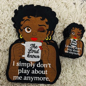 Könnte beinhalten: Gestickter Aufnäher mit einer Cartoon-Frau mit schwarzen Haaren, goldenen Ohrringen und rotem Lippenstift, die eine Tasse hält. Auf der Tasse steht "The Lord knows". Der Text "I simply don't play about me anymore" steht darunter. Eine kleinere Version ist ebenfalls vorhanden.