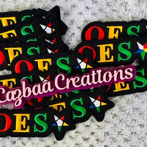 May include: Several black embroidered patches displaying the letters "OES" in red, yellow, and green. Some patches have a star design with colourful accents. A pink and purple "Cazbaa Creations" label is also visible.