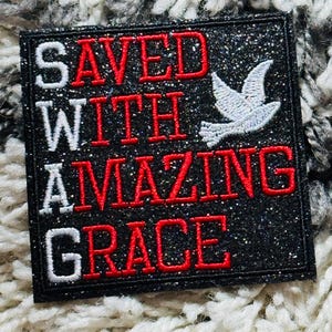 May include: Square black patch with glitter, featuring the words "SAVED WITH AMAZING GRACE" in red and white lettering. A white dove is embroidered on the right side. The patch has a black border.