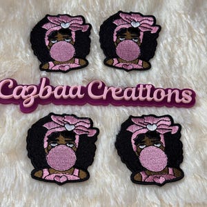 Peut inclure: Quatre écussons brodés représentant une fille aux cheveux noirs, avec un nœud rose et une bulle de chewing-gum. Les écussons ont une bordure noire et sont sur une surface blanche et duveteuse. Le texte "Cazbaa Creations" est également visible.