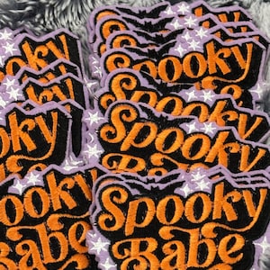 May include: Embroidered patches with the words "Spooky Babe" in orange script on a black background. The patches have a purple border with white stars and a bat design. Halloween-themed.