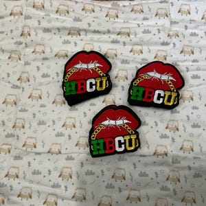 May include: Three embroidered patches featuring red lips with gold chain detailing and the letters HBCU in green, black, and gold. The patches are on a white fabric with a repeating owl pattern.
