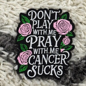 May include: Black embroidered patch with the text "DON'T PLAY WITH ME PRAY WITH ME CANCER SUCKS" surrounded by pink roses and green leaves. The patch is on a textured, light-colored surface.