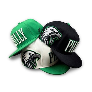 May include: Three baseball caps stacked together. One is bright green with white text reading PHILLY. One is white with a black and white eagle graphic. One is black with a white and green eagle graphic and white text reading PHILLY.