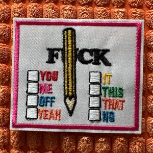 Puede incluir: Parche bordado con la palabra "F*ck" y un diseño de lápiz. El parche incluye casillas de verificación con las palabras "You, Me, Off, Yeah" y "It, This, That, No". El parche tiene un borde rosa.