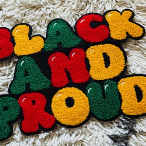 Può includere: Un arazzo tessile trapuntato che riporta le parole BLACK AND PROUD in un carattere grassetto e arrotondato. Le lettere sono testurizzate e colorate in rosso, giallo e verde vivaci, con riflessi bianchi su uno sfondo nero uniforme.