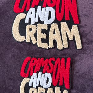 Może przedstawiać: Dwie haftowane naszywki z napisem "CRIMSON AND CREAM" pogrubioną, blokową czcionką. Słowo "CRIMSON" jest czerwone, "AND" jest białe, a "CREAM" jest w kolorze beżowym. Naszywki znajdują się na ciemnofioletowym tle.
