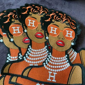 Puede incluir: Tres parches bordados con la ilustración estilizada de una persona con cabello oscuro corto, que lleva gafas de sol redondas naranjas con una letra H blanca en cada lente, un collar de perlas blancas de varias hebras y una blusa naranja con una H blanca.
