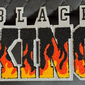 May include: A silver-bordered patch with the words "BLACK KING" in a varsity-style font. The letters are black with a flame design in orange and yellow. The patch is likely for clothing or accessories.