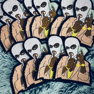 May include: Embroidered patches featuring a figure wearing a white ski mask, a tan suit, and gold jewelry. The figure holds a wad of cash and gestures with a finger to the lips, indicating silence. The patches have a black border.