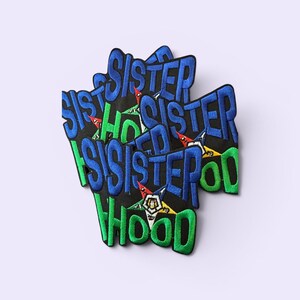 May include: Embroidered patches with the words "Sisterhood" in blue and green. The patches have a black background and feature a star design with red, yellow, and green accents. The patches are stacked on top of each other.