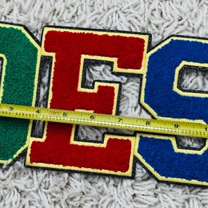 May include: A set of three varsity-style letters spelling OES, featuring a green O, red E, and blue S. Each letter has a yellow border and a black outline. A yellow measuring tape is placed across the letters, showing a width of 11 inches.