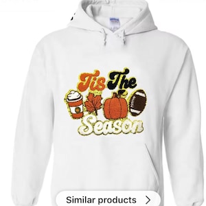 Peut inclure: Sweat-shirt à capuche blanc avec un motif sur le thème de l'automne. Le design comprend une tasse de café, une feuille d'érable, une citrouille et un ballon de football, avec le texte "Tis The Season" en orange, noir et or.