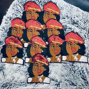 Puede incluir: Un conjunto de parches bordados que representan a una mujer con piel oscura, labios rojos y un boina roja. La boina tiene el texto "LOUIS VUITTON" en dorado. La mujer se toca la cara con una mano. Los parches están sobre una superficie gris texturizada.