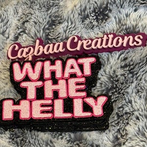 May include: Two layered, decorative signs with text on a textured, grey background. The top sign reads "Cazbaa Creations" in pink script. The bottom sign says "WHAT THE HELLY" in pink block letters with a black glitter outline.