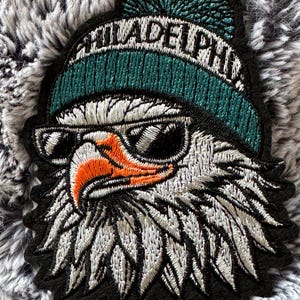 May include: Embroidered patch of an eagle wearing sunglasses and a teal beanie with "PHILADELPHIA" in white. The eagle has an orange beak and is set against a black border. Ideal for clothing.