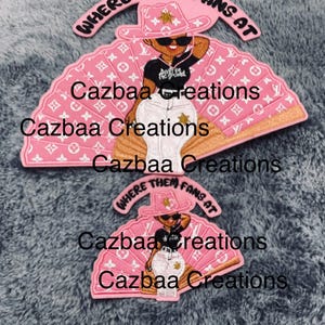 Puede incluir: Accesorio rosa en forma de abanico con un diseño de vaquera de dibujos animados. La vaquera lleva un sombrero rosa y gafas de sol, con el texto "WHERE THEM FANS AT". El abanico tiene un patrón repetitivo y el texto "Cazbaa Creations".