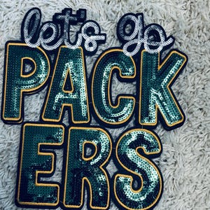 Op de afbeelding: Een pailletten patch met de woorden "let's go PACKERS" in groen, goud en zilver. De letters zijn zwart omlijnd en gevuld met groene pailletten. De woorden "let's go" zijn in zilveren pailletten.