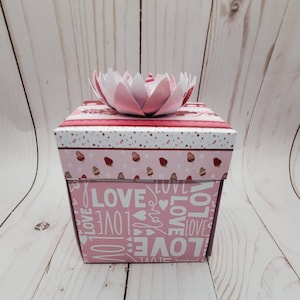 May include: A pink and white explosion box with a pink paper flower on top. The box is decorated with a pattern of strawberries and the word "love" repeated many times.