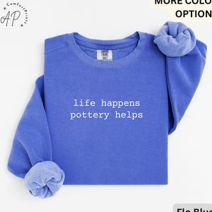 May include: A blue crewneck sweatshirt with the text "life happens pottery helps" in white. The sweatshirt is a solid blue color and has a folded sleeve. The image also includes the words "MORE COLOR OPTIONS" and "Flo Blue".