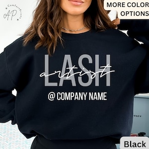 Peut inclure: Sweat-shirt noir avec l'inscription "LASH artist @ COMPANY NAME" en gris et blanc. Le sweat-shirt a un col rond et des manches longues. Les mots "MORE COLOR OPTIONS" sont en haut à droite. Le mot "Black" est en bas à droite.