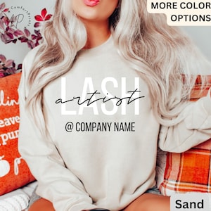 Peut inclure: Un sweat-shirt blanc avec le texte "LASH artist" et "@COMPANY NAME" en noir. Le sweat-shirt est porté par une personne aux longs cheveux blonds. L'arrière-plan est une image floue d'une couverture à carreaux et d'un oreiller.