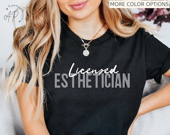 Op de afbeelding: Zwart T-shirt met de tekst "Licensed Esthetician" in wit en grijs. Het shirt heeft ook de tekst "Color - Black" onderaan. Het T-shirt heeft een ronde hals en wordt gedragen.