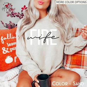 May include: A beige sweatshirt with the text "FIRE wife" in black lettering. The sweatshirt is being worn by a person sitting on a sofa with a red and white checked throw pillow.