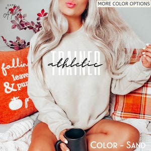 May include: A beige sweatshirt with the text "TRAINER athletic" in black lettering. The sweatshirt is being worn by a person sitting on a sofa with a cushion that says "fallin leave & pur".