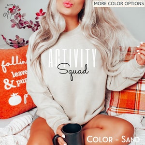 May include: A beige sweatshirt with the text "ACTIVITY Squad" printed in black. The sweatshirt is being worn by a person sitting on a sofa with a cushion that says "fallin leave & pur".