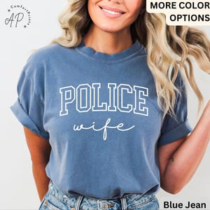 May include: Blue t-shirt with the words "POLICE wife" in white block letters and script. The shirt has rolled-up sleeves and is made of a soft material. The shirt is a blue jean color.