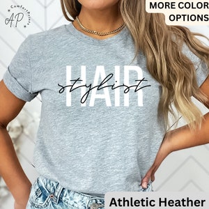 K&ouml;nnte beinhalten: Ein graues T-Shirt mit dem Text "HAIR stylist" in wei&szlig;en Buchstaben. Das Shirt wird von einer Person mit langen blonden Haaren getragen.
