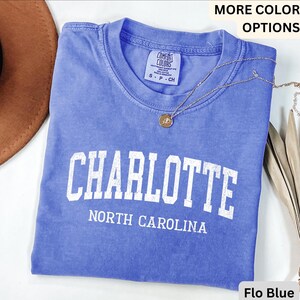 Op de afbeelding: Een blauw t-shirt met het woord "CHARLOTTE" in grote witte letters en "NORTH CAROLINA" eronder. Het shirt heeft een ronde hals en korte mouwen. Een gouden ketting met een hanger is over het shirt gedrapeerd.
