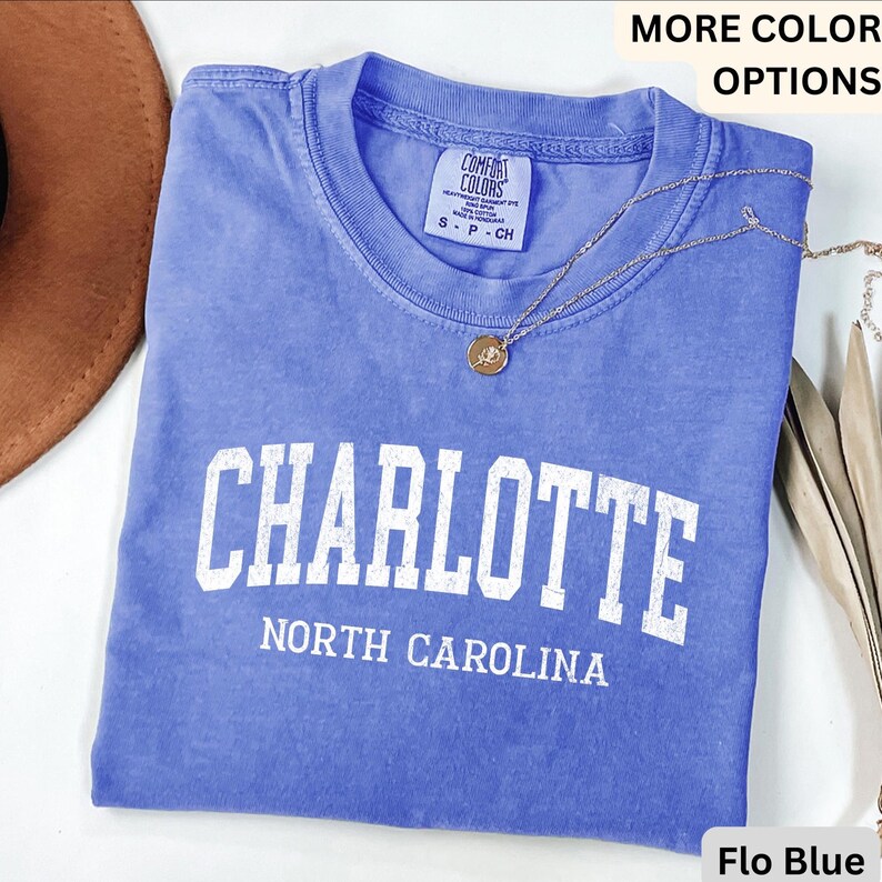 Op de afbeelding: Een blauw t-shirt met het woord "CHARLOTTE" in grote witte letters en "NORTH CAROLINA" eronder. Het shirt heeft een ronde hals en korte mouwen. Een gouden ketting met een hanger is over het shirt gedrapeerd.
